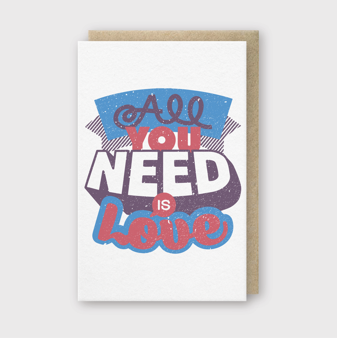 The "All You Need is Love Greeting Card" showcases vibrant, bold lettering in shades of blue, purple, pink, and red with a distressed texture. This handcrafted card is designed in Seattle and includes an eco-friendly recycled Kraft envelope set against a white background, combining charm with environmental consciousness.