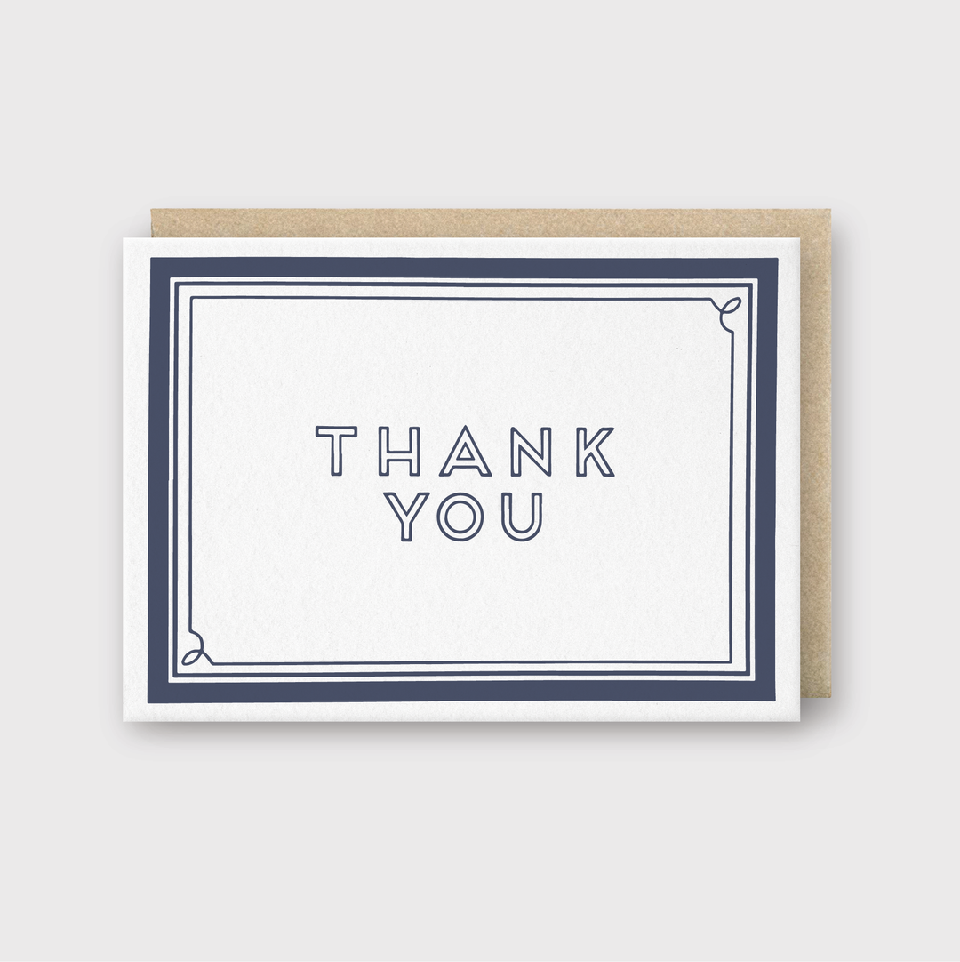 The Thank You Navy Frame Greeting Card offers a simple yet elegant design featuring "Thank You" in bold, capital letters centered within a thin navy blue frame. Using premium letterpress techniques, this handcrafted card is paired with a neutral brown envelope.