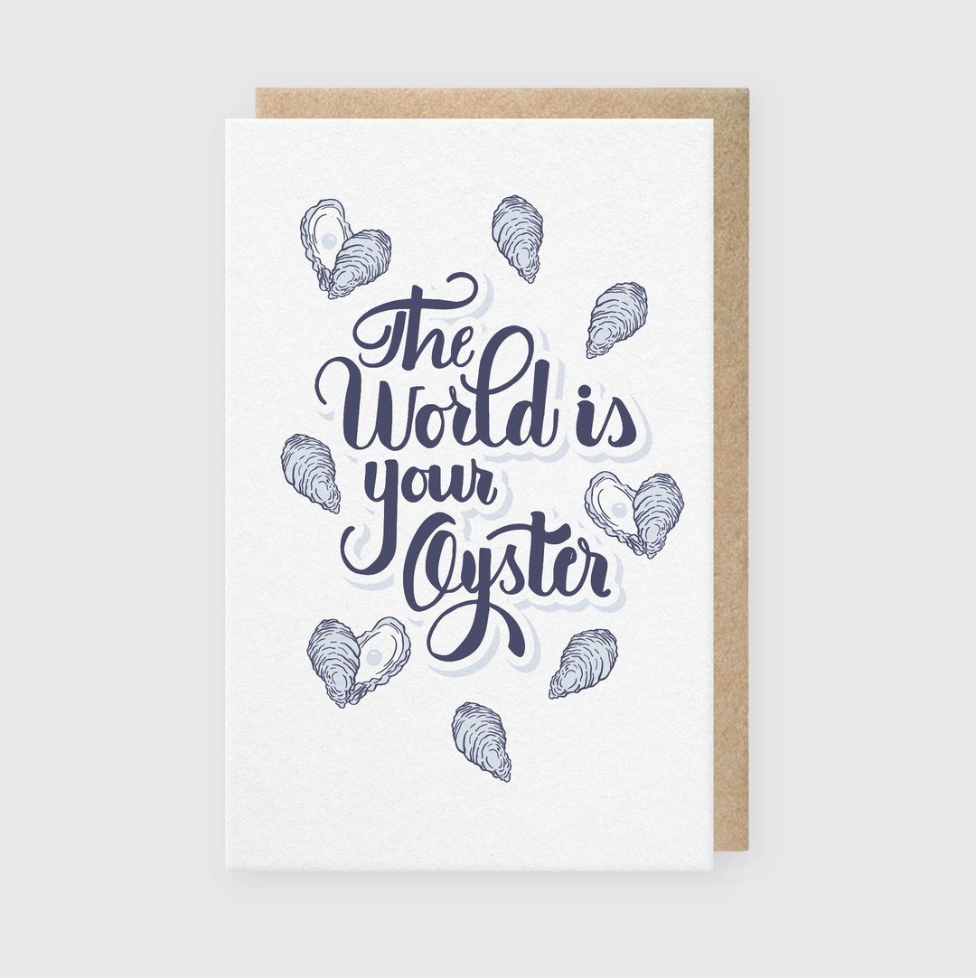 Introducing the "The World Is Your Oyster Greeting Card," this letterpress card features artistic blue lettering of the inspirational phrase and is adorned with oyster shell illustrations on a white background. It comes paired with a brown kraft envelope.