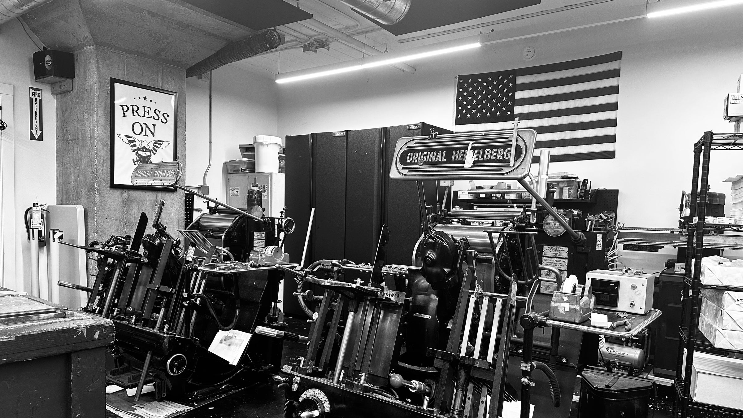 The black and white image captures a nostalgic ambiance of a print shop, showcasing vintage Heidelberg printing presses as the focal point. An American flag adds a touch of historical pride on the wall, alongside an inspiring "Press On" poster that embodies perseverance and dedication to craft. The workspace is characterized by its industrial elements—metallic surfaces, sturdy machinery—and neatly organized shelves that underline the orderly nature of such an establishment.