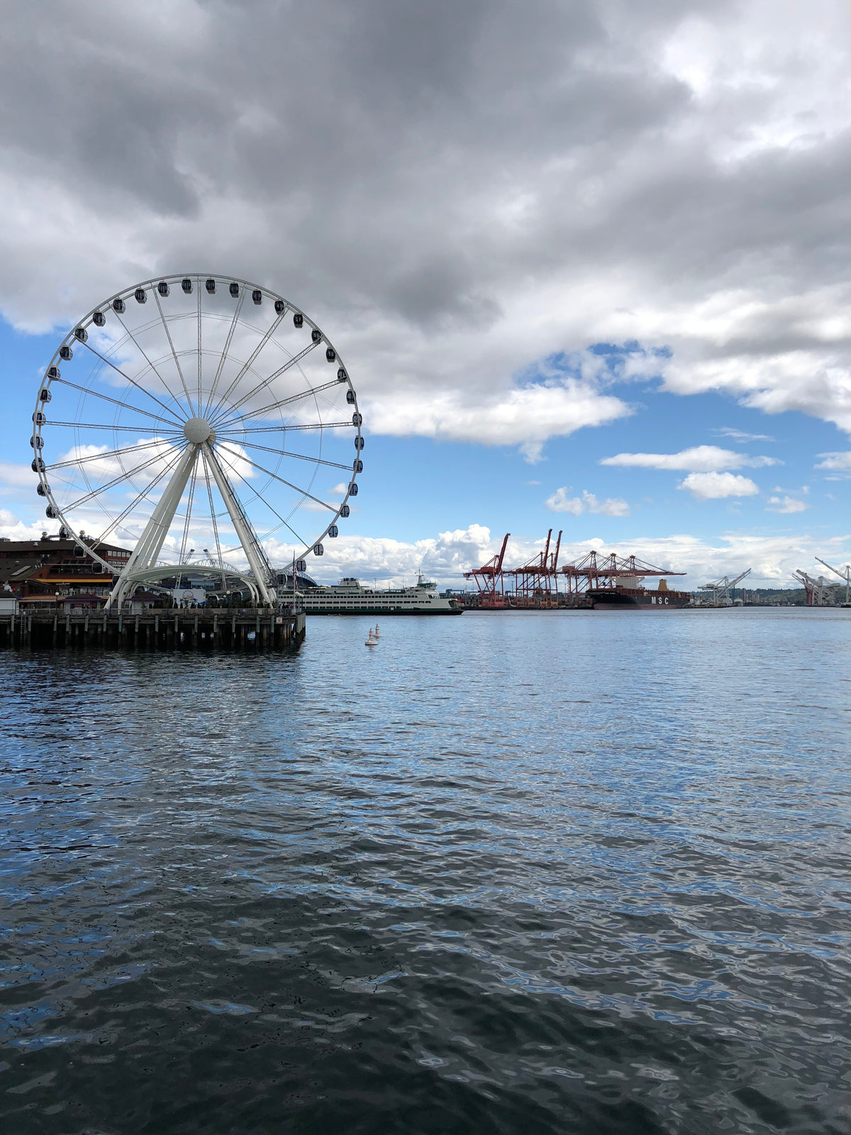 A large Ferris wheel stands on a waterfront, with cranes and industrial structures in the background. The sky is partly cloudy, and the water reflects the scene, creating a serene coastal atmosphere.