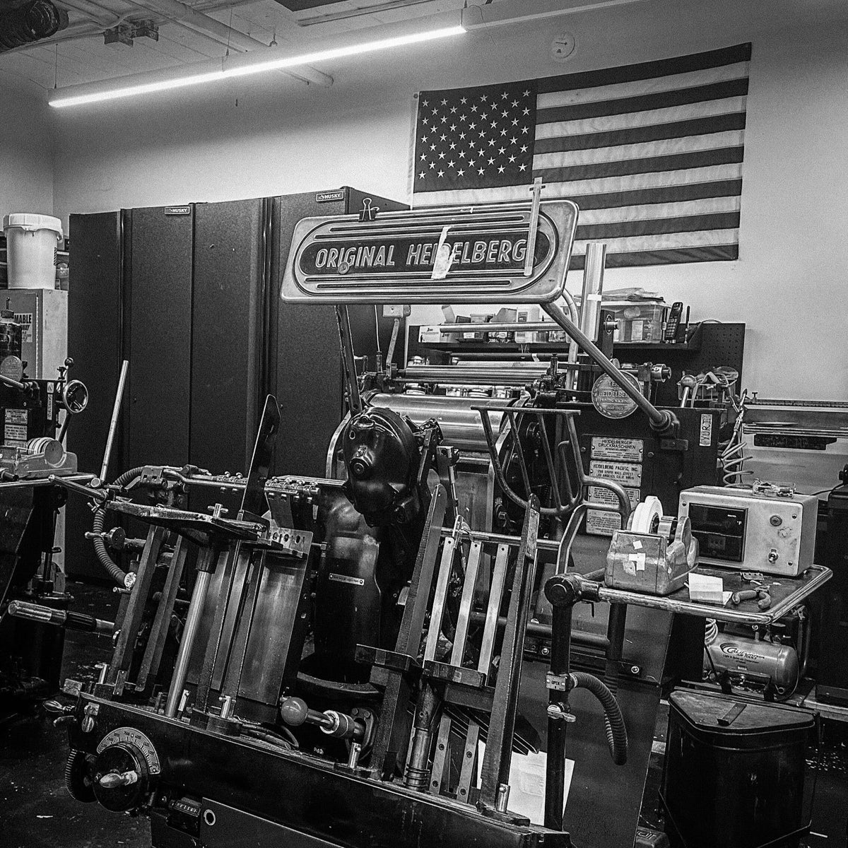 The image depicts a classic printing press, known as the "Original Heidelberg," situated in a workshop. The setting is marked by an assortment of tools and materials essential for traditional printmaking. In the backdrop, an American flag adds a touch of character to the scene, highlighting its locale. The black and white aesthetic lends an authentic vintage feel to the photograph, capturing the essence of historical craftsmanship.