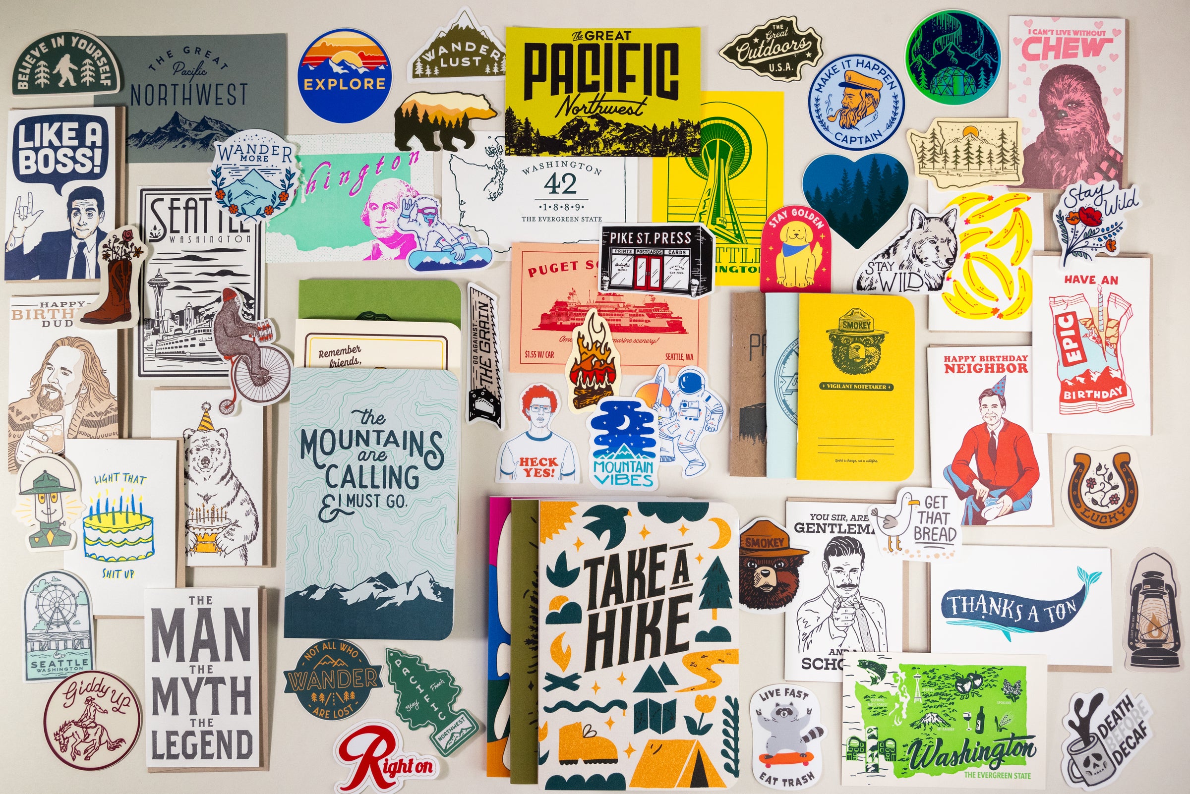 This is a vibrant assortment showcasing a variety of stickers, posters, and artwork. The designs prominently feature travel themes and include motivational quotes like "Take a Hike." Emphasizing Northwest motifs, the collection displays images of mountains, trees, and wildlife against a crisp white background.