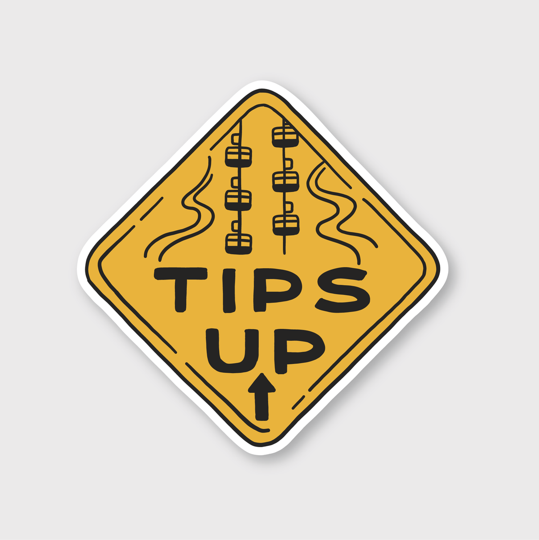 The sticker is diamond-shaped and yellow, featuring bold black letters that say "TIPS UP." Above the text are illustrations of three chairlifts with wavy lines, all made from high-quality vinyl. A small black arrow points upwards, emphasizing the artistic design.