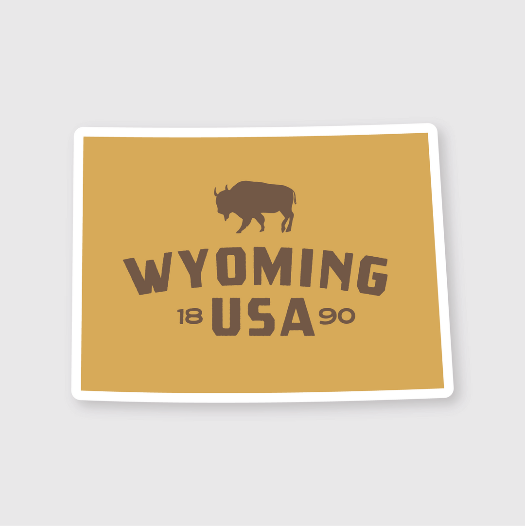 The Wyoming Simple State Sticker by Pike Street Press is displayed on a golden brown postcard. It features a die-cut vinyl sticker with a bison silhouette and bold text reading "WYOMING USA," with the year creatively split around the letters.