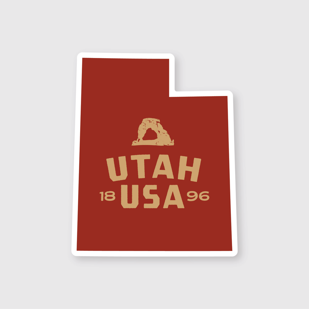 The Utah Simple State Sticker is a high-quality die-cut vinyl sticker in the shape of Utah. It features a striking red background with a cream illustration of an arch, and bold cream text that reads "Utah USA 1896," celebrating the state’s rich history and beauty.