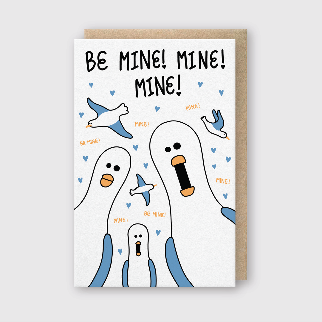 The "Be Mine Mine Mine Greeting Card" features animated cartoon seagulls enthusiastically saying "Be Mine! Mine! Mine!" against a white background decorated with small blue hearts. The card is handcrafted in Seattle and shows these lively seagulls in various poses. It comes with a recycled Kraft envelope.