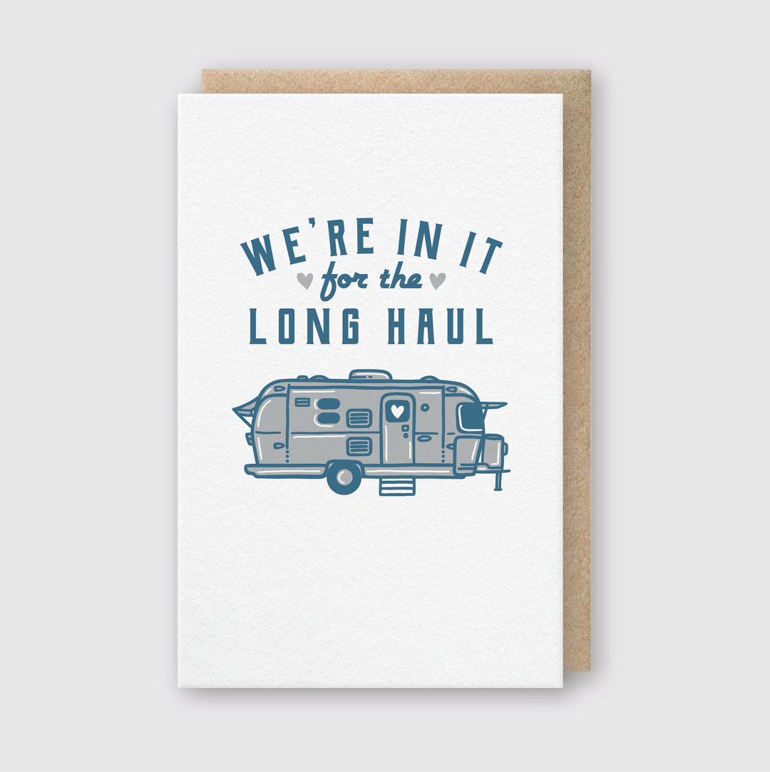 The "In It For The Long Haul Greeting Card" features a charming vintage camper illustration with the phrase "WE'RE IN IT for the LONG HAUL" in blue lettering. The design is adorned with small heart motifs and is set against a light gray background. A classic brown envelope accompanies this delightful card.