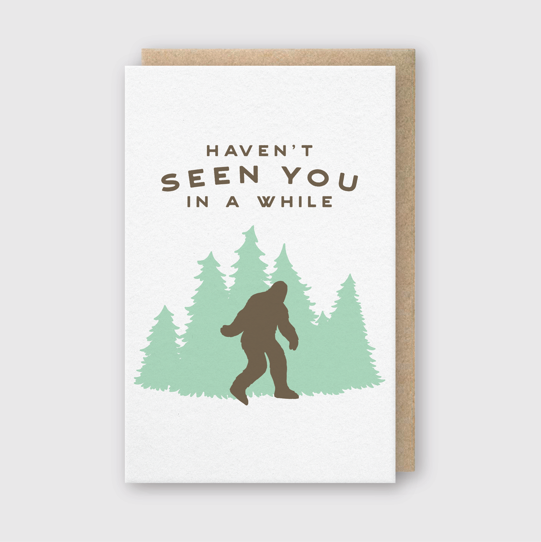 The "Haven't Seen You In A While Greeting Card" is a handcrafted card featuring a silhouette of Bigfoot walking past lush green pine trees. It is printed on FSC certified paper and includes the text, "Haven’t seen you in a while," at the top.