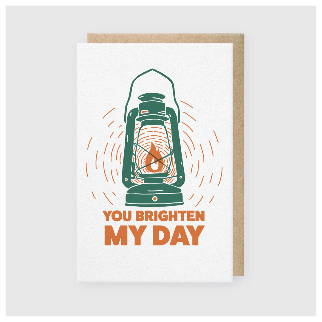 The "You Brighten My Day Lantern Greeting Card" features a vintage lantern design that casts light, accompanied by the message "You Brighten My Day" in bold orange letters. It's printed with soy-based inks on a light gray background.