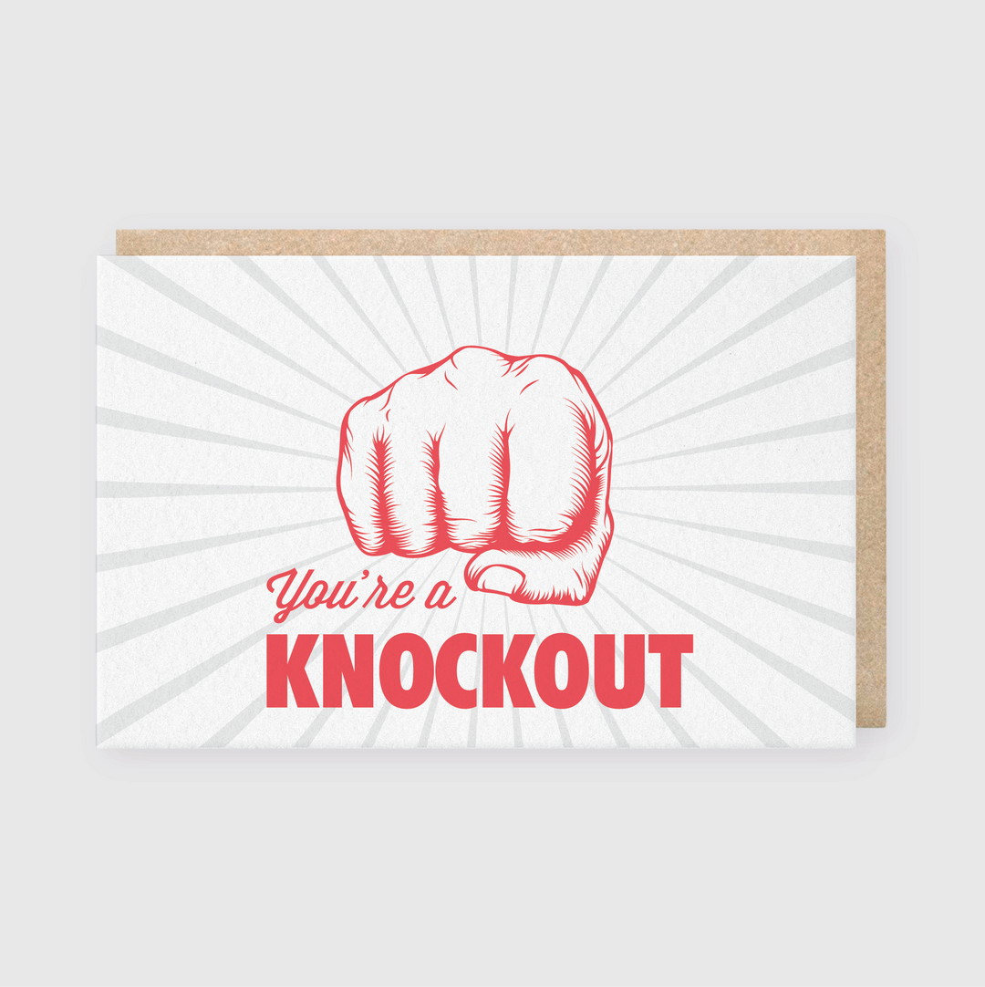 Introducing the "You're A Knockout Greeting Card," a handcrafted masterpiece from Pike Street Press. This card features a bold red illustration of a fist, enhanced by dynamic sunburst lines and encouraging text, delivering an impactful message: "You're a KNOCKOUT!