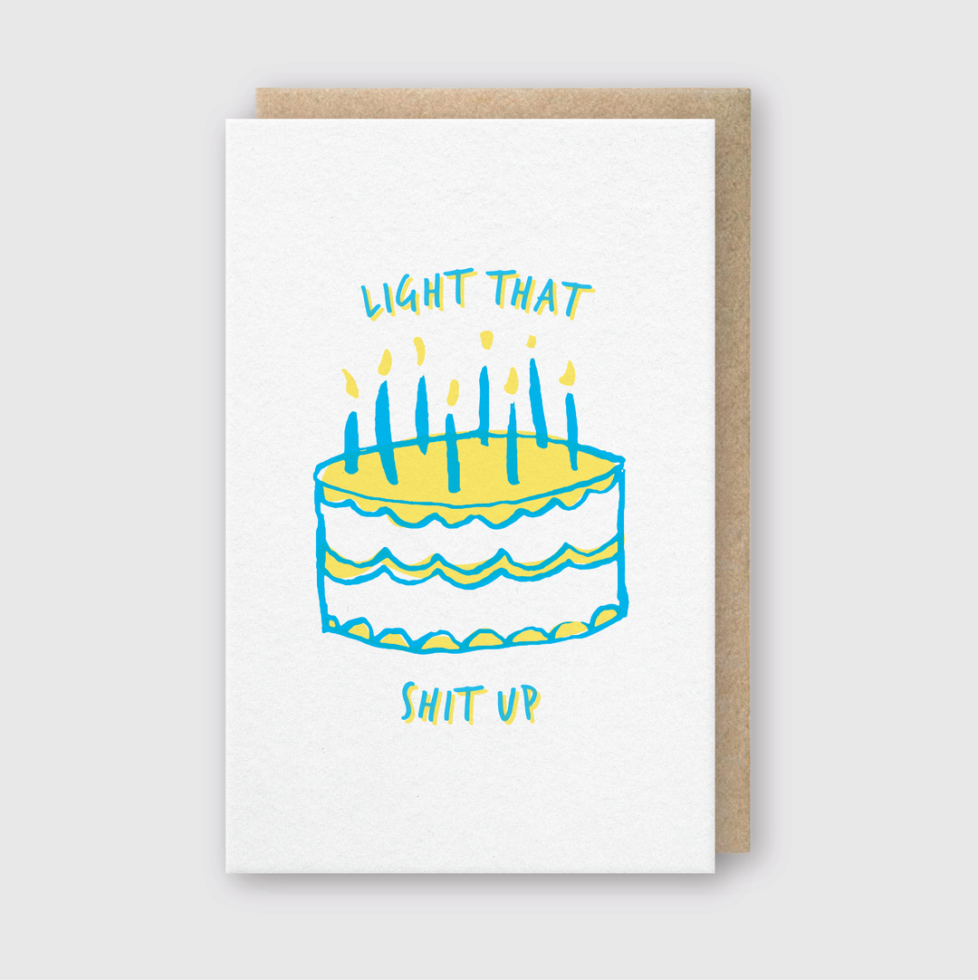 The Light That Up Greeting Card showcases a cake with lit candles on a white background. The card humorously features the text "LIGHT THAT" at the top and "SHIT UP" at the bottom. It comes with a recycled kraft envelope, making it an eco-friendly choice for birthday greetings.