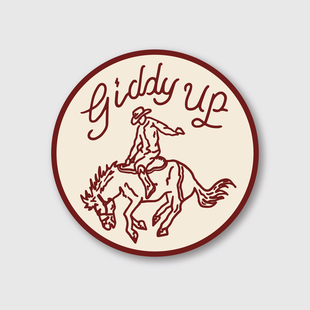 The "Giddy Up Sticker" from Pike Street Press is a die-cut vinyl piece that exudes a handcrafted, vintage appeal. It features an illustration of a person riding a bucking horse with the words "Giddy Up" elegantly scripted above, all encapsulated in a circular design with rustic charm.