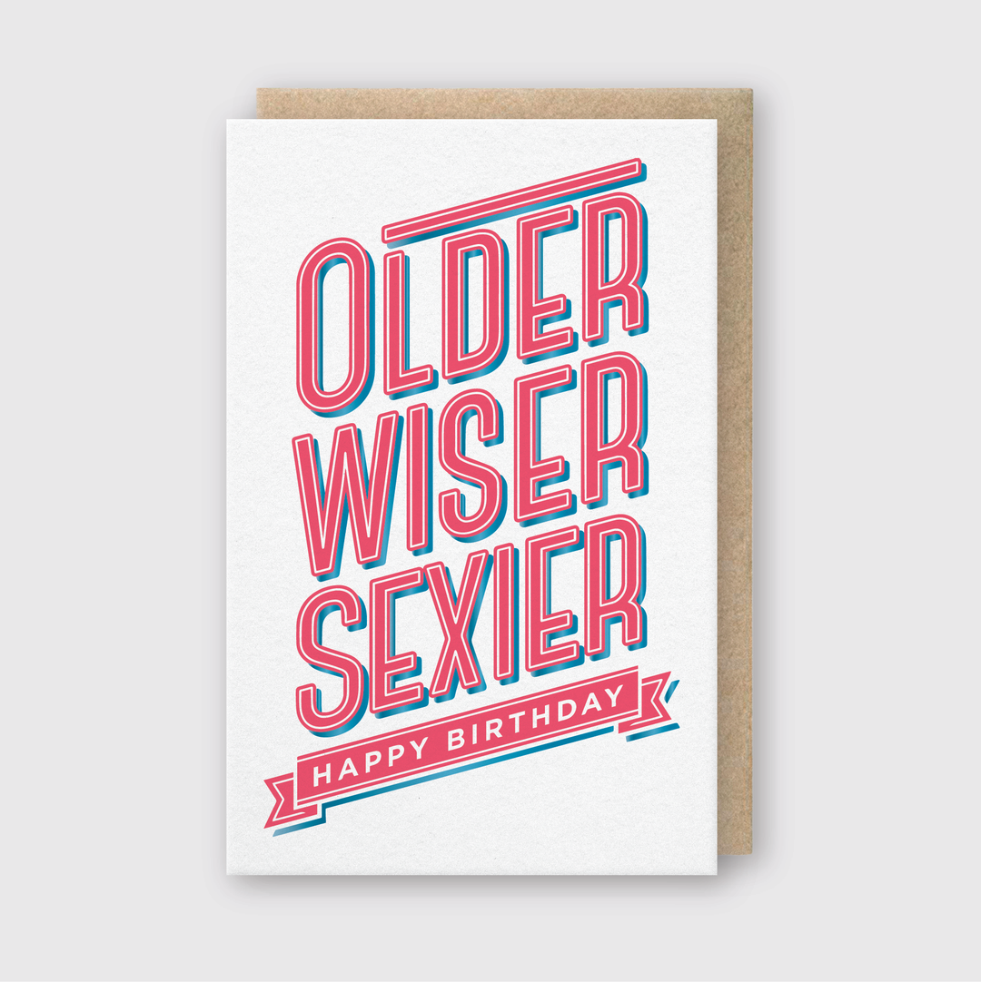 The "Older Wiser Sexier" greeting card is a handcrafted piece featuring bold, layered red and blue text. It presents the words "Older Wiser Sexier" prominently above a red banner that reads "Happy Birthday." The card is complemented by a brown envelope and displayed on a light gray background, highlighting its design elements.