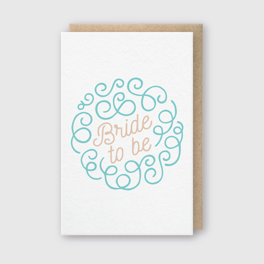 The greeting card features the phrase "Bride to be" elegantly centered within a decorative teal scroll design. It comes with a simple brown envelope and is photographed on a light gray background.