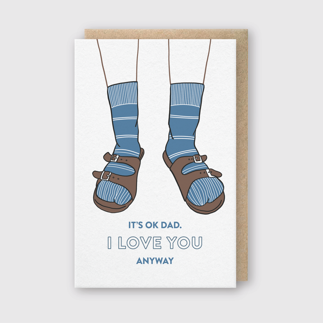 The Dad Birkenstocks Greeting Card features an illustration of legs in blue socks and brown sandals, with the text "It's OK Dad. I love you anyway." It comes with a brown envelope and is perfect for Father's Day. This card is handcrafted in Seattle, adding an extra touch of love.