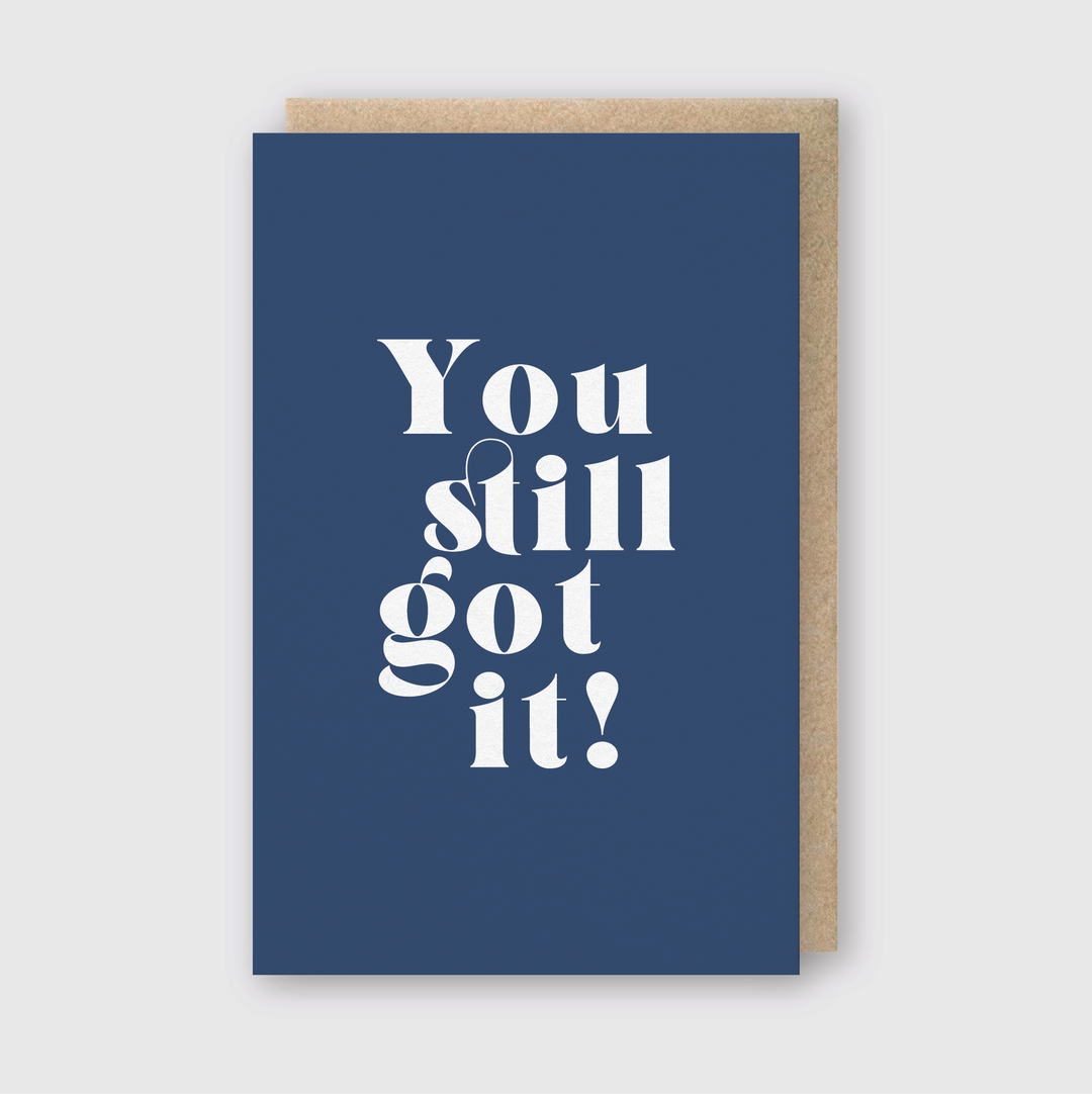 The "You Still Got It Greeting Card" features a vibrant blue cover adorned with bold, playful white text that exclaims, "You still got it!" This card is meticulously handcrafted in Seattle using environmentally friendly FSC Certified Paper. The design is showcased slightly open, highlighting its natural brown backing set against a subtle light gray background.