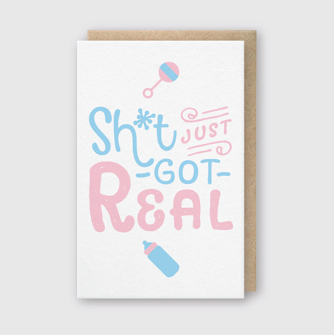 Introducing the "Sh*t Just Got Real" Greeting Card, a playful design in pink and blue fonts for new parents. Handcrafted in Seattle, this card features a white background adorned with delightful graphics of a baby bottle and rattle, making it perfect for celebrating this exciting chapter.