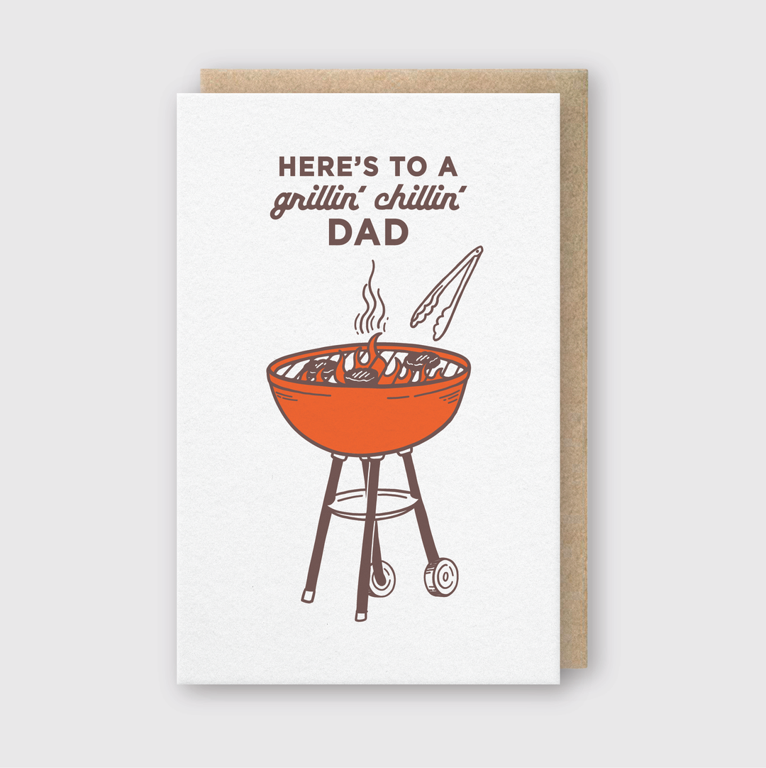 The Grillin' Chillin' Dad Greeting Card features a red charcoal grill with steaming food and tongs on the cover. The card's text reads: "Here's to a grillin' chillin' dad." It's an ideal choice for fathers who enjoy barbecuing, contrasting beautifully against a plain background.