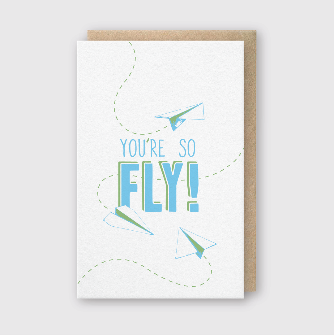 The "You're So Fly Greeting Card" is a whimsical card featuring playful blue and green lettering with paper airplane designs. Handcrafted in Seattle, it comes with a recycled kraft envelope, making it an eco-friendly choice for any occasion.