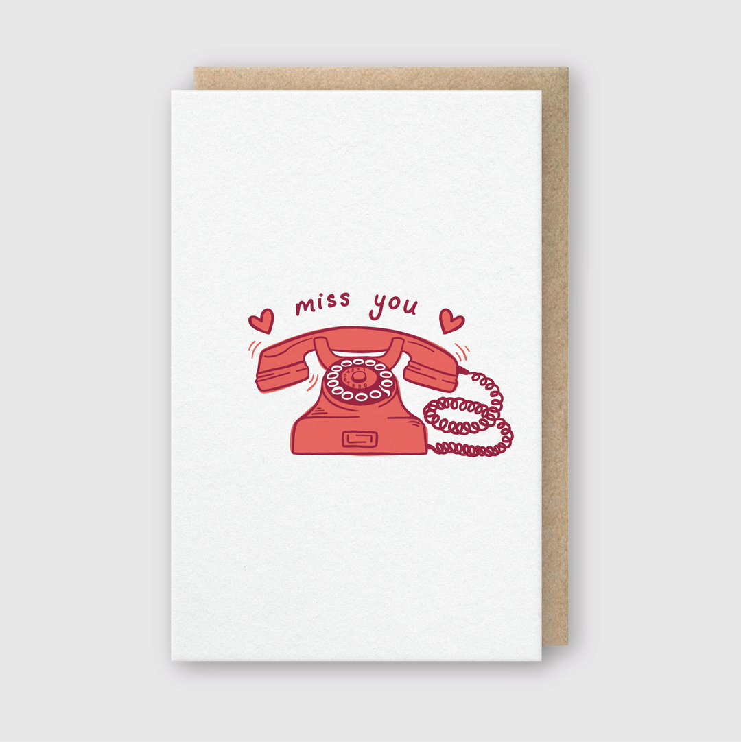 The "Miss You Phone Greeting Card" features a charming illustration of a red retro rotary phone with a curly cord. Above the phone, the words "miss you" are elegantly inscribed and surrounded by small red hearts. This card is hand printed in Seattle on FSC Certified Paper and is accompanied by a light brown envelope, providing an elegant presentation.