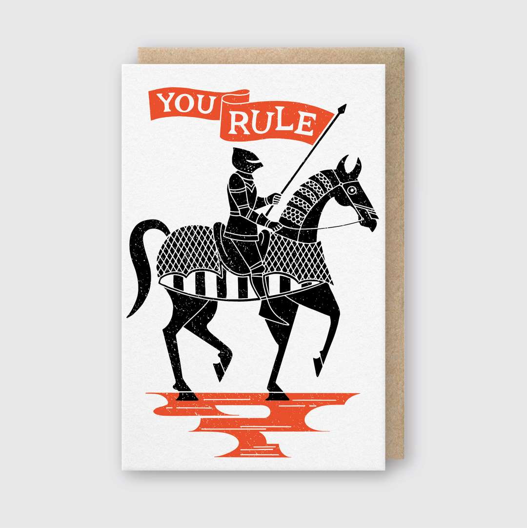 The "You Rule Greeting Card" features a medieval knight in armor, gallantly riding a horse with a lance. An orange banner above the knight declares "YOU RULE," while the striking black and orange design contrasts against a white backdrop, all elegantly encased in a classic brown envelope.