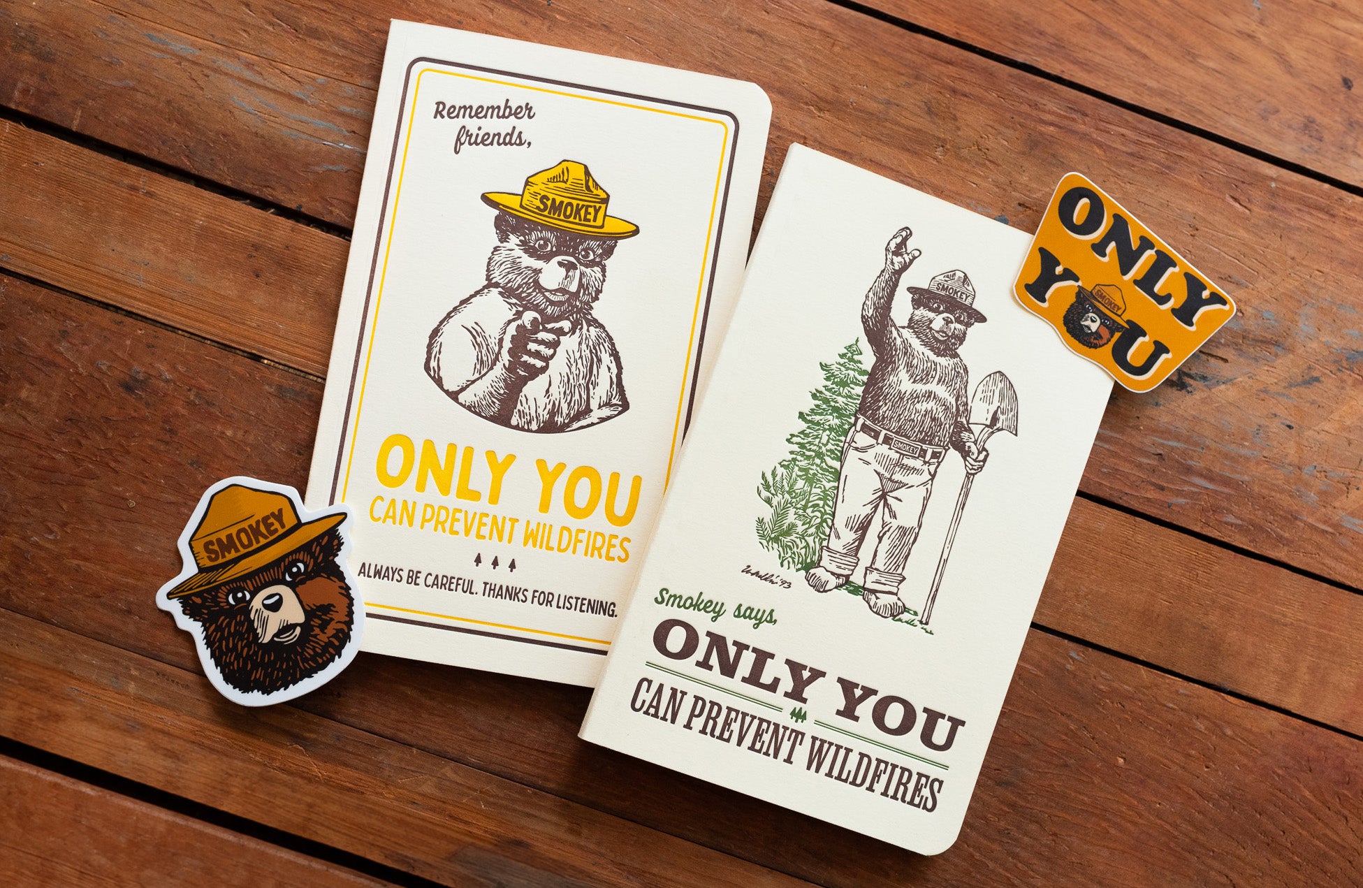 Two booklets and stickers featuring Smokey Bear are placed on a wooden surface. The booklets carry the message "Only you can prevent wildfires" and include illustrations of Smokey Bear wearing ranger hats.