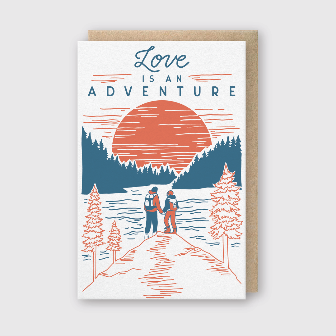 The "Love Is An Adventure Hikers Greeting Card" presents a charming illustration of a couple holding hands as they admire a picturesque lake and forest landscape, highlighted by a large setting sun. The phrase "Love is an Adventure" complements the card's vibrant red, blue, and white color palette.