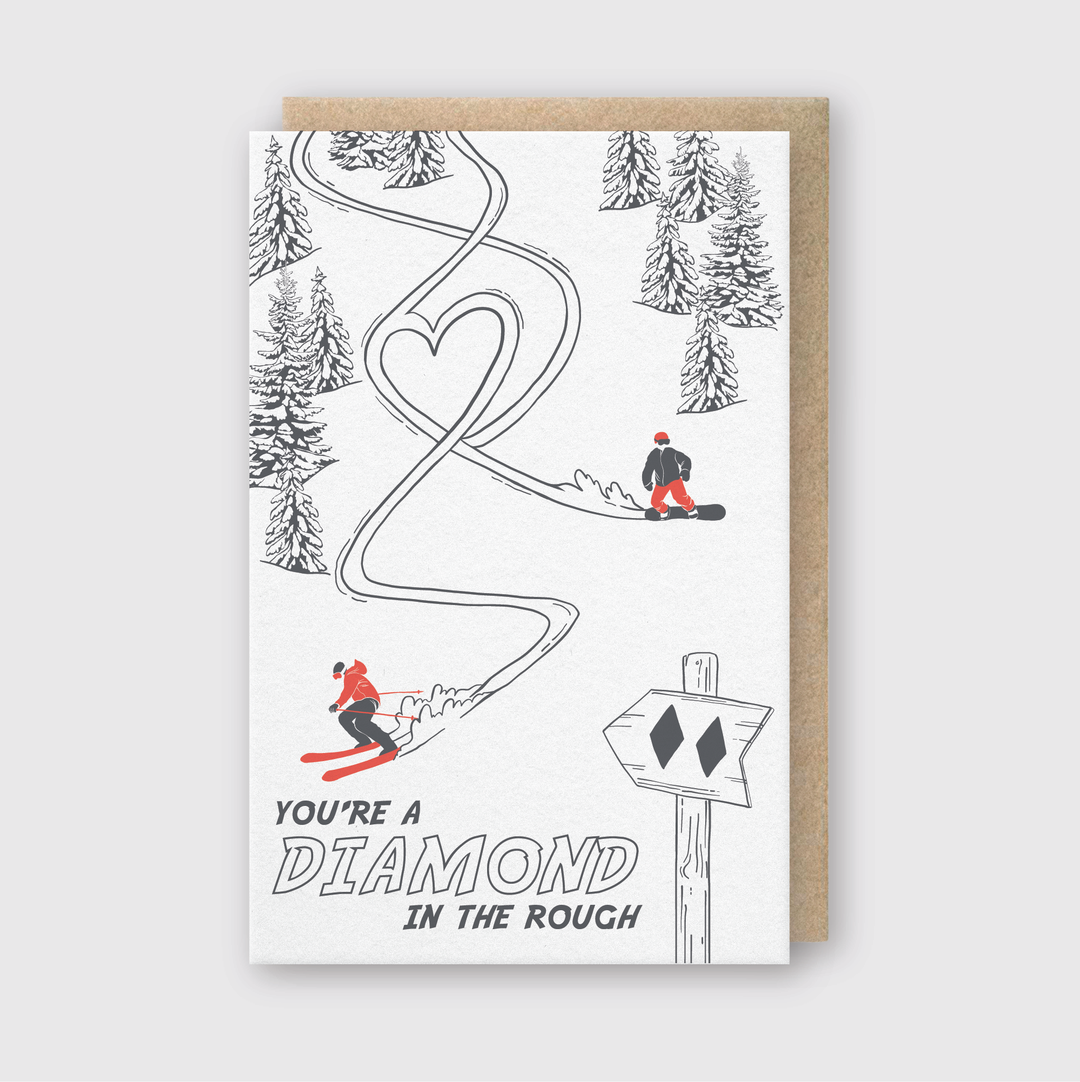 The Diamond in the Rough Greeting Card is a beautifully crafted card featuring an illustration of two skiers on a snowy slope, leaving heart-shaped trails amidst pine trees. A sign indicating a double black diamond run is also depicted. The card includes the message, "You're a Diamond in the Rough," and is printed on FSC Certified Paper, reflecting its environmentally friendly production. This handcrafted card originates from Seattle and offers a charming way to convey your sentiments.