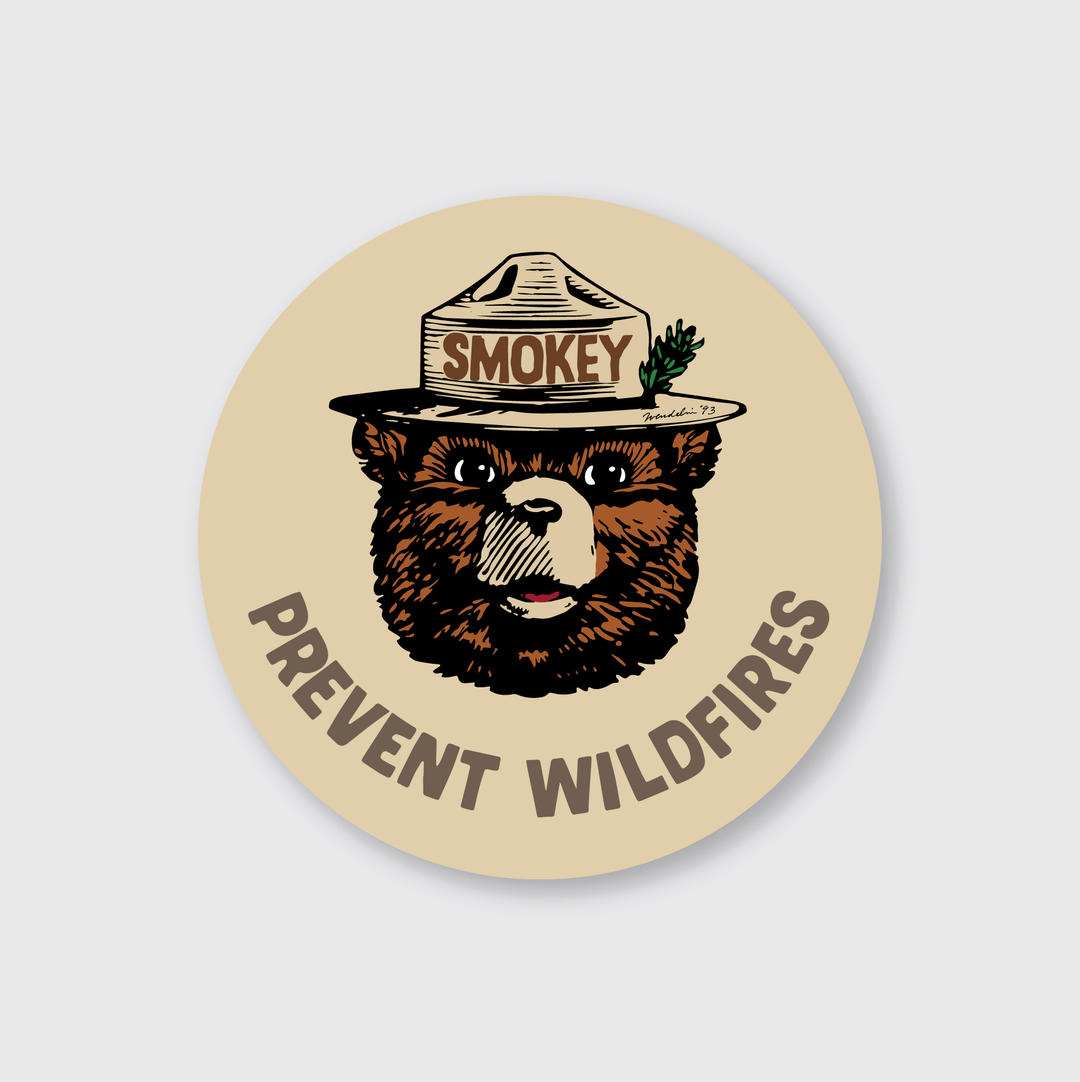 The Smokey Bear Prevent Wildfires Badge Sticker features an illustration of Smokey Bear wearing a park ranger hat with "Smokey" inscribed on it. Below the image, bold lettering on a beige background reads "Prevent Wildfires," enhancing the bear's serious expression to underscore its authoritative message.