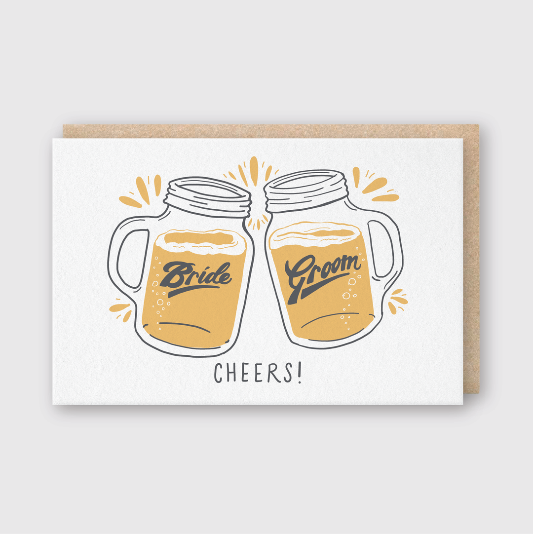 The Cheers Beers Bride & Groom Greeting Card is a handcrafted card featuring an illustration of two beer mugs clinking together, with "Bride" and "Groom" inscribed on them, and the word "CHEERS!" underneath. It comes from Seattle and is packaged in a brown envelope.