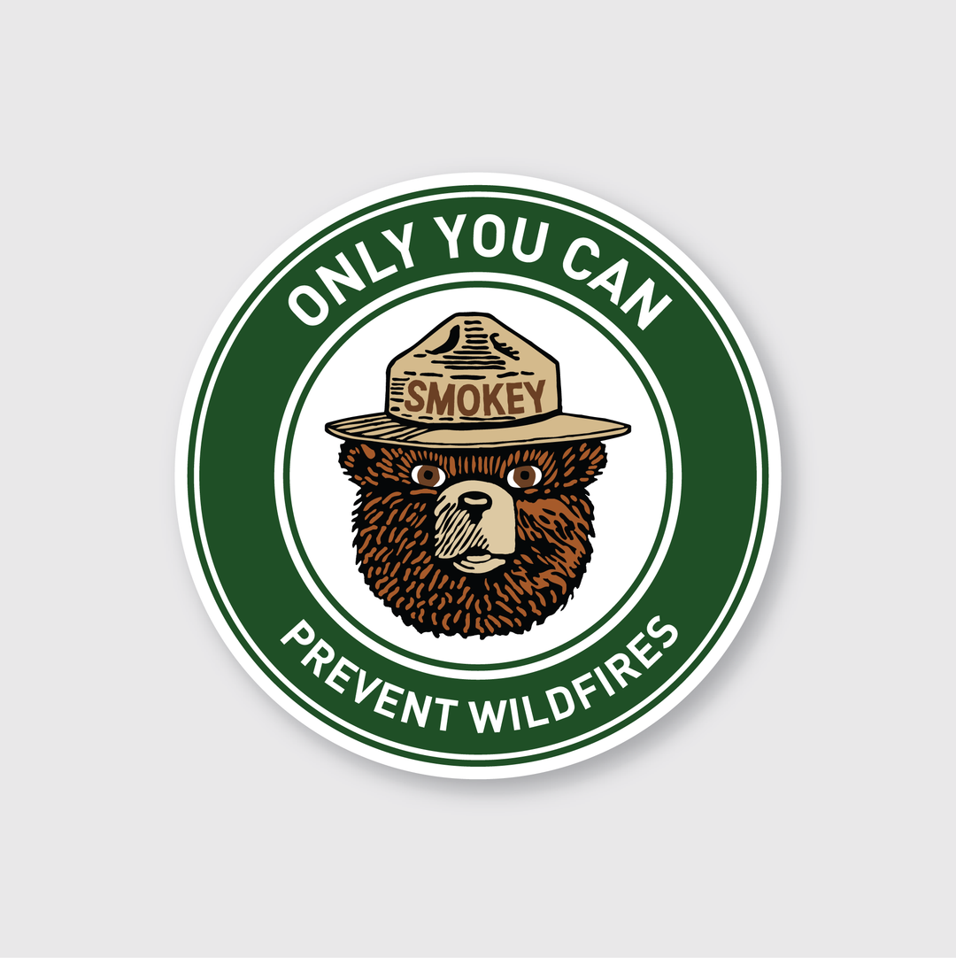 The Smokey Bear Slogan Badge Sticker features a circular badge design of Smokey Bear wearing a ranger hat with the label "Smokey." The slogan "Only You Can Prevent Wildfires" encircles the image on a green background. This die-cut vinyl sticker is weather-resistant, designed for long-lasting durability and awareness.