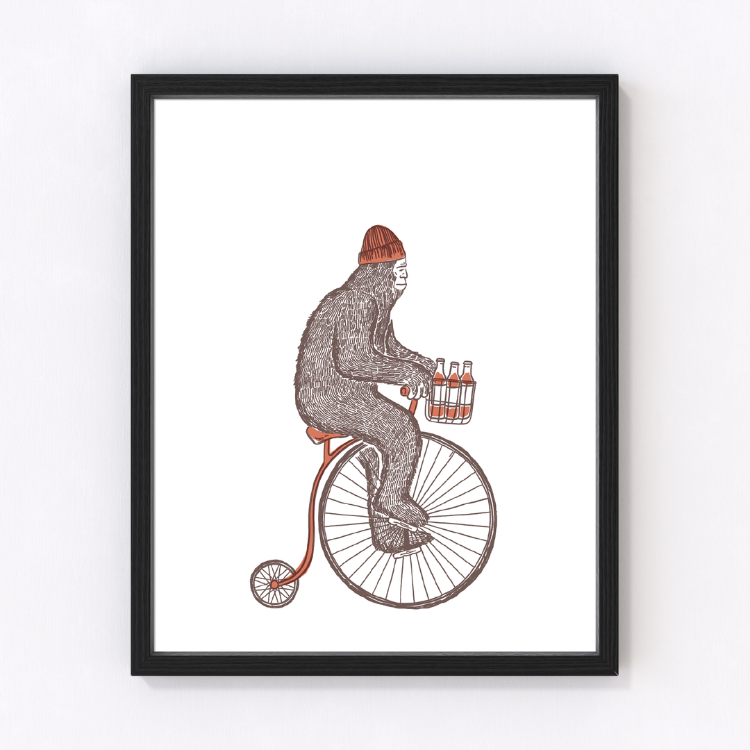 Bigfoot in a red beanie rides a vintage penny-farthing while carrying a six-pack, framed in black and hung on a white wall.