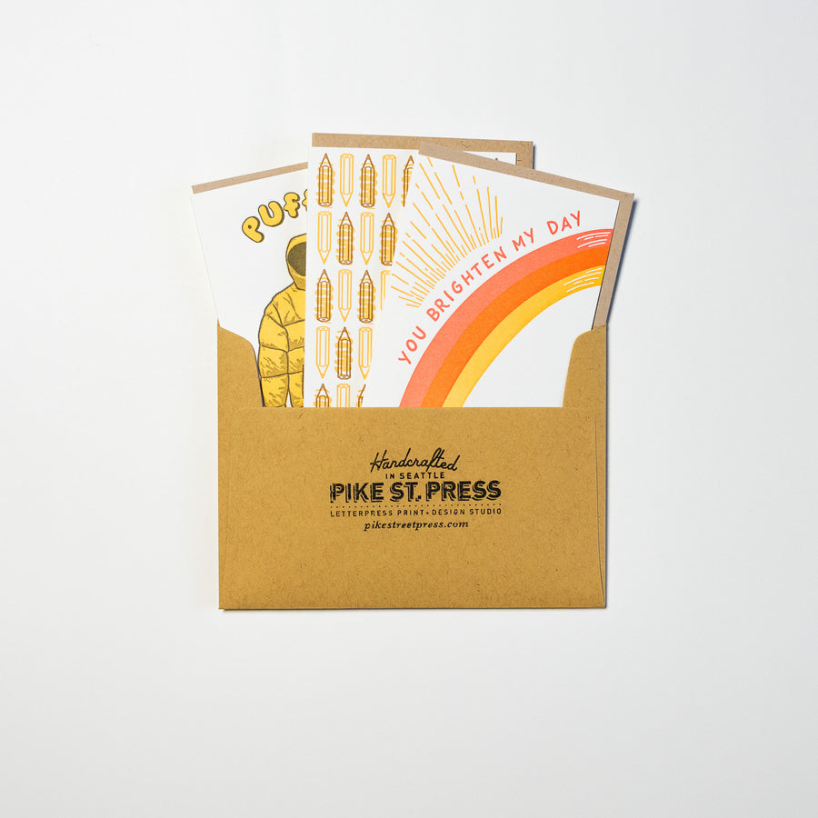 A brown envelope labeled "Pike Street Press" unveils two vibrant cards from their Monthly Subscription Box. One card showcases a rainbow with the message "YOU BRIGHTEN MY DAY," while the other features illustrated corndogs, both exuding color and playfulness.