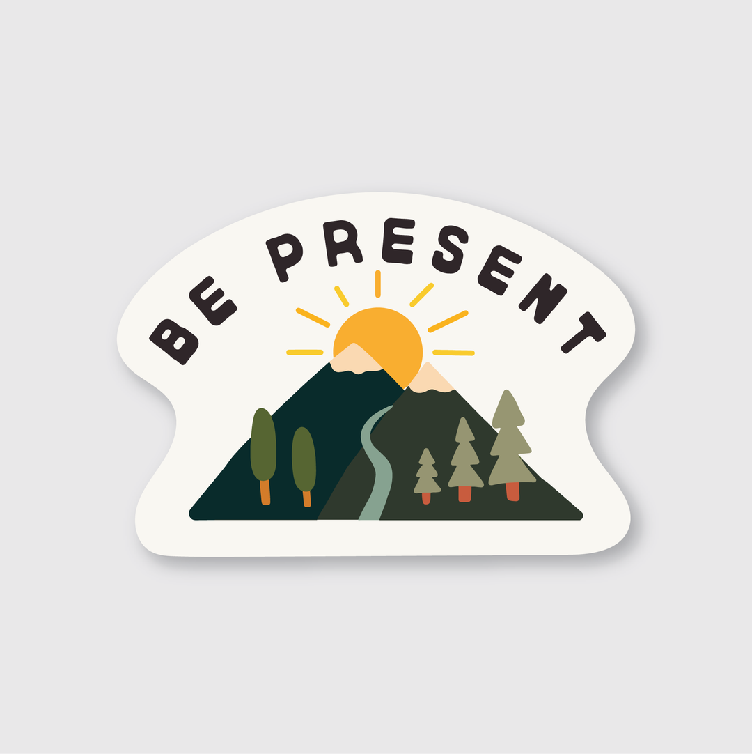 The "Be Present Mountains Sticker" by Pike Street Press features an illustration of a sunrise over a mountain with trees in the foreground and the bold text "Be Present" above. This premium die-cut vinyl sticker is weather-resistant with a laminated matte finish, making it ideal for both indoor and outdoor inspiration.