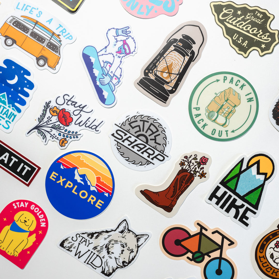 A collection of vibrant outdoor-themed stickers is displayed on a white surface. The stickers feature illustrations such as a van, lantern, wolf, bicycle, hiking boot, and mountains. Accompanying these designs are phrases like "Stay Wild," "Explore," and "Life's a Trip.